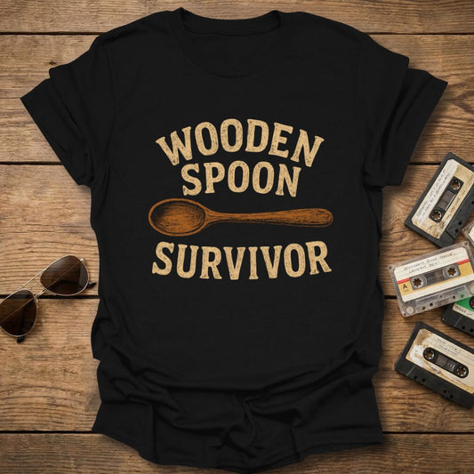 Black t-shirt with 'Wooden Spoon Survivor' text on a wooden surface with vintage items.