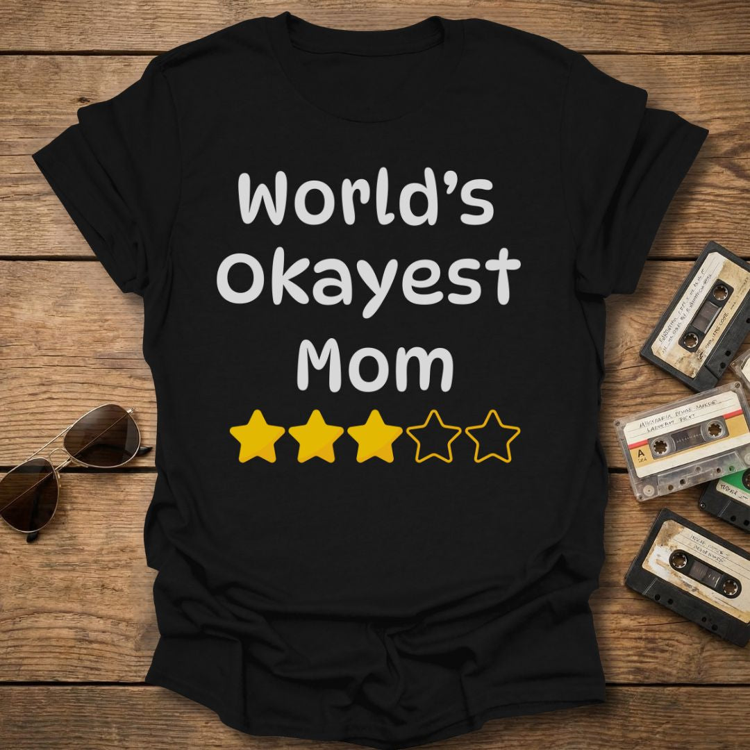 Black t-shirt with 'World's Okayest Mom' text and 3 star rating on a wooden surface with vintage items.