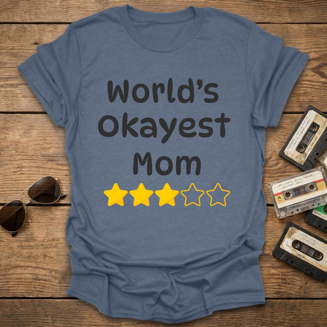 Indigo t-shirt with 'World's Okayest Mom' text and 3 star ratings on a wooden surface with vintage cassettes and sunglasses.
