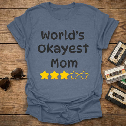 Indigo t-shirt with 'World's Okayest Mom' text and 3 star ratings on a wooden surface with vintage cassettes and sunglasses.