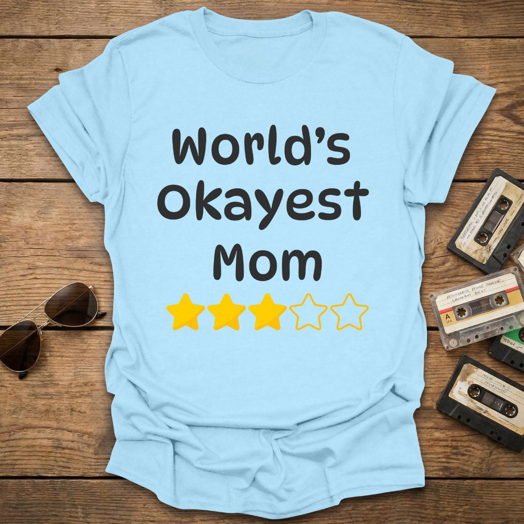Light blue t-shirt with 'World's Okayest Mom' text and 3 star rating on a wooden surface with vintage items.