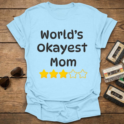 Light blue t-shirt with 'World's Okayest Mom' text and 3 star rating on a wooden surface with vintage items.