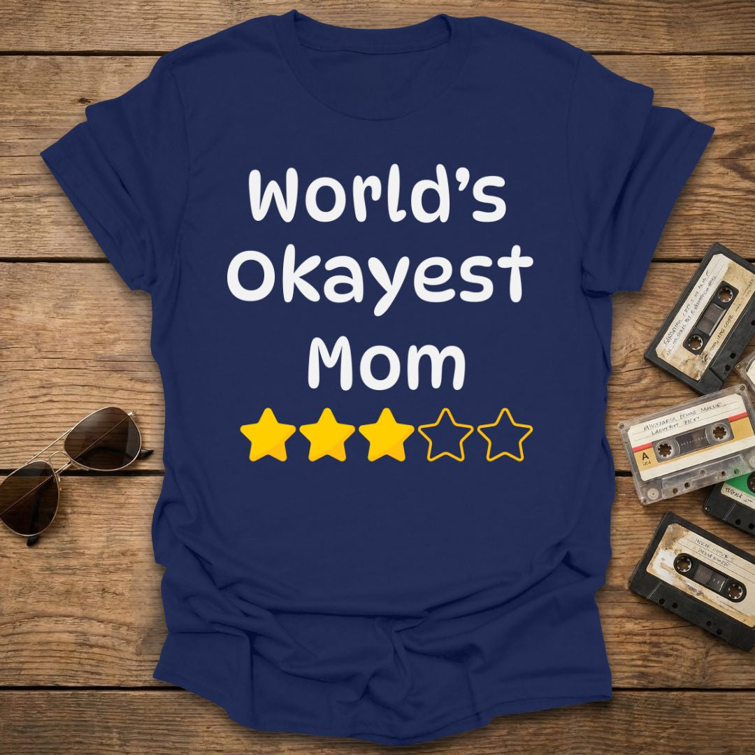 Navy blue t-shirt with 'World's Okayest Mom' text and star ratings on a wooden surface with vintage cassettes and sunglasses.