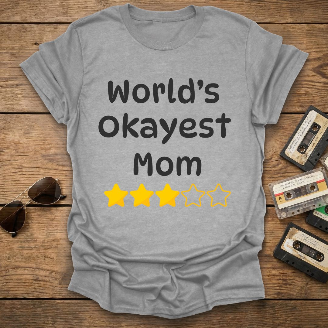 Gray t-shirt with 'World's Okayest Mom' text and 3 star ratings on a wooden surface with vintage cassettes and sunglasses.