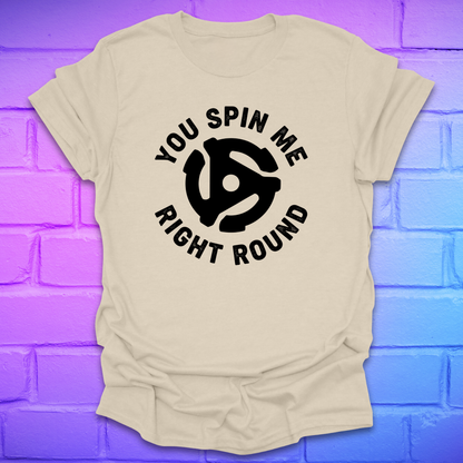 Beige t-shirt with black graphic and text ,'you spin me right round' on a purple brick wall background